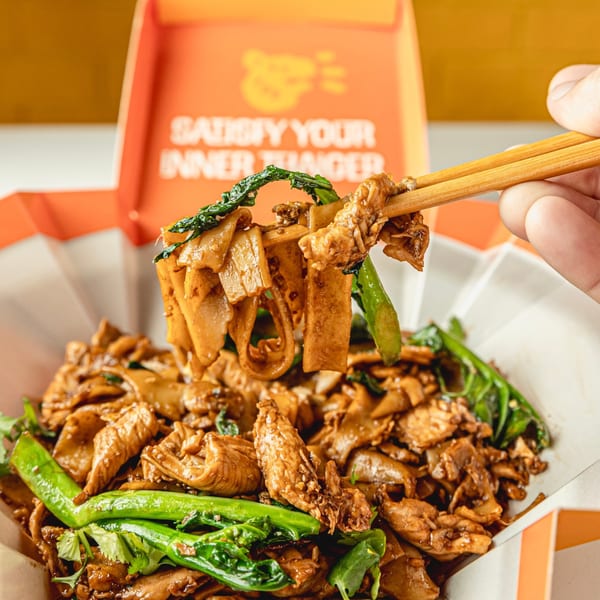 Chopsticks lifting wide noodles, chicken, and green vegetables from a Thai Tail takeout box.