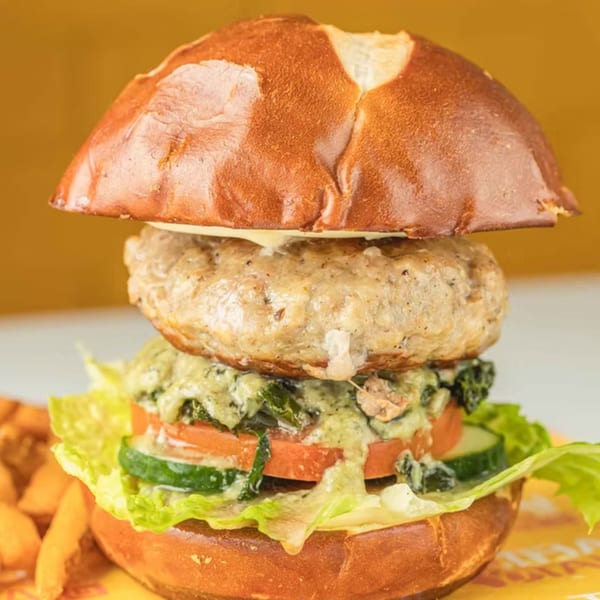 A Thai Tail burger with a pretzel bun, patty, cheese, lettuce, tomato, cucumber, and green sauce.