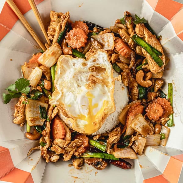 Thai chicken stir-fry with carrots, cashews, and a fried egg over rice in a takeout container.