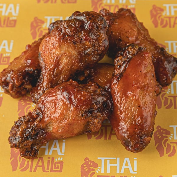 A pile of glossy, dark brown Thai chicken wings on a yellow background with the Thai Tail logo.